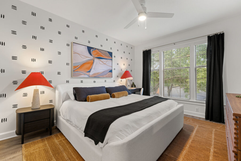 A bedroom with a white upholstered bed, black-and-orange accents, and abstract wall art, centered on a patterned area rug in front of a window with black curtains.