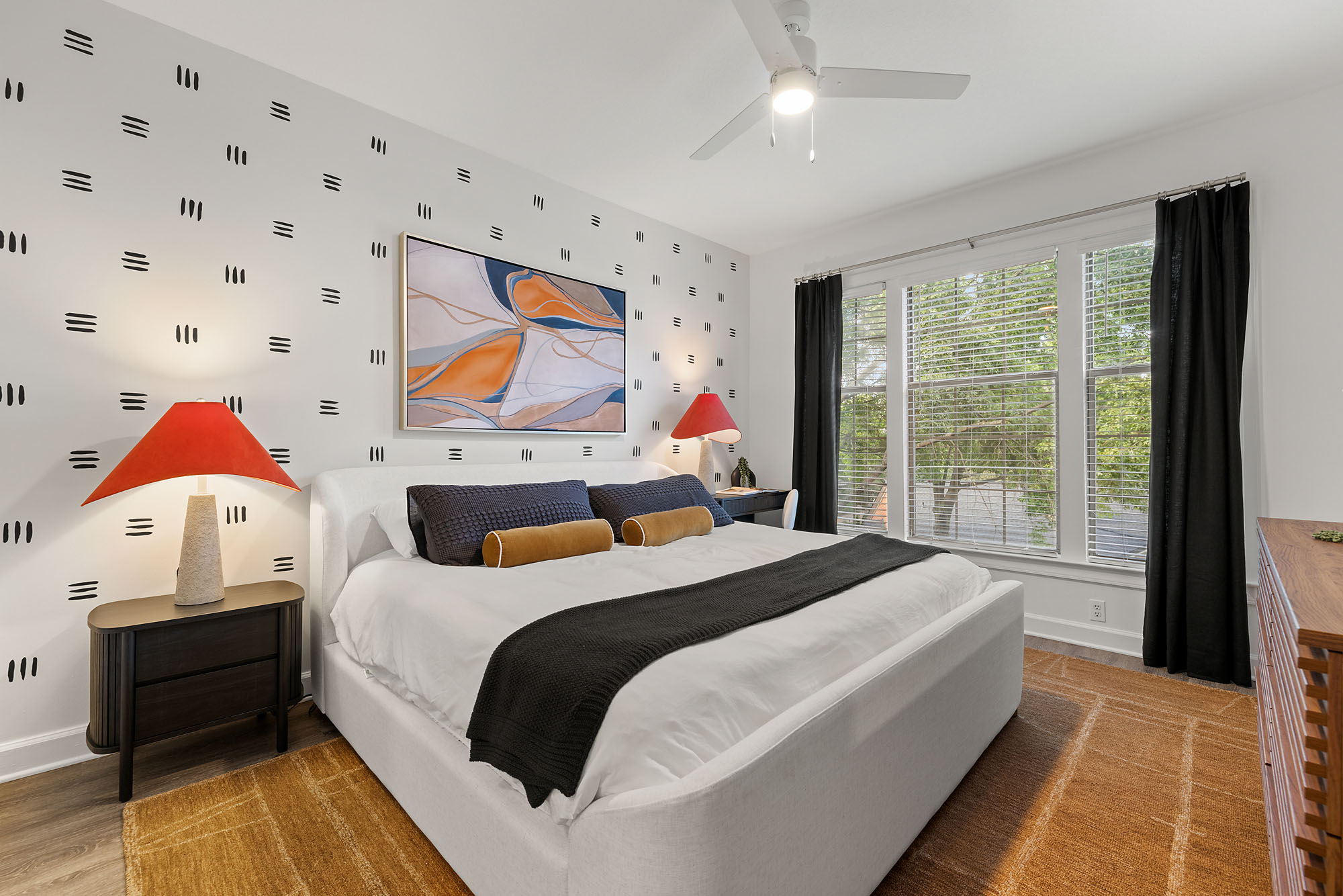 A bedroom with a white upholstered bed, black-and-orange accents, and abstract wall art, centered on a patterned area rug in front of a window with black curtains.