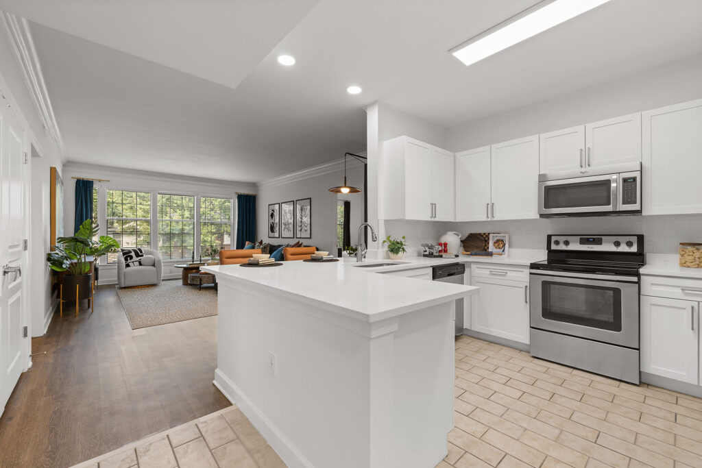 A modern kitchen with a white island and stainless steel appliances flows into an open-concept living area featuring an armchair, a leather sofa, and a large window.