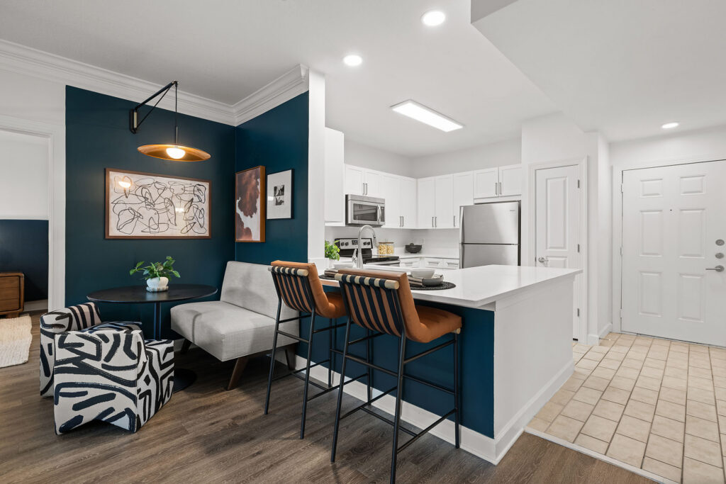 A dining area with a round table, patterned chairs, and a bench, next to a kitchen with white cabinets, stainless steel appliances, and a breakfast bar with two stools.