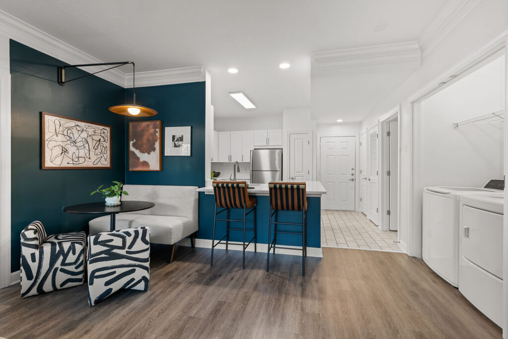 A dining area with a small round table, a bench, and two patterned armchairs sits next to a kitchen island with two bar stools, featuring a dark blue accent wall with framed artwork.