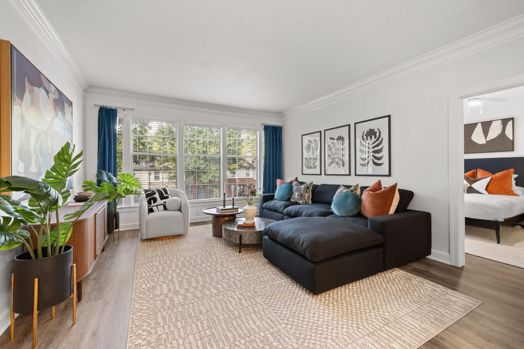 A living room featuring a black sectional sofa, a light gray armchair, two coffee tables, and a large potted plant, with a view into a bedroom beyond.