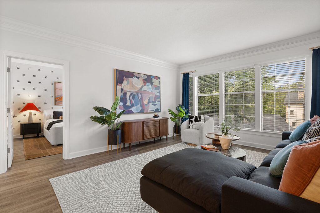 A living room featuring a dark sofa, a wooden console table with an abstract painting above it, an armchair by the windows, and a doorway leading to a bedroom with a black lamp.