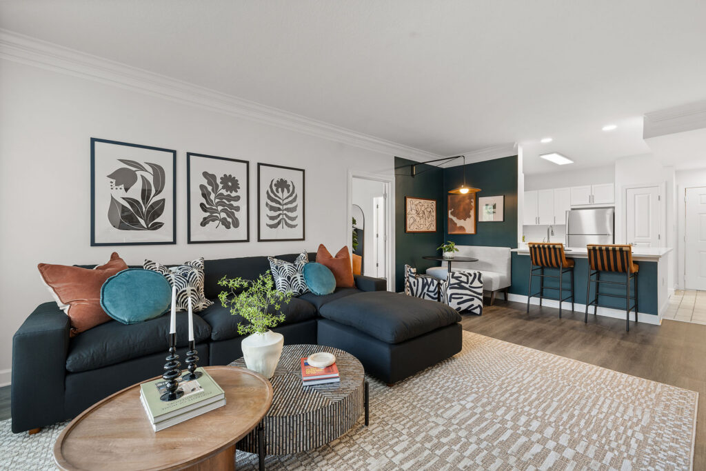 A living room featuring a dark sofa with decorative pillows, two circular coffee tables, and a dining area with patterned chairs and a kitchen island in the background.